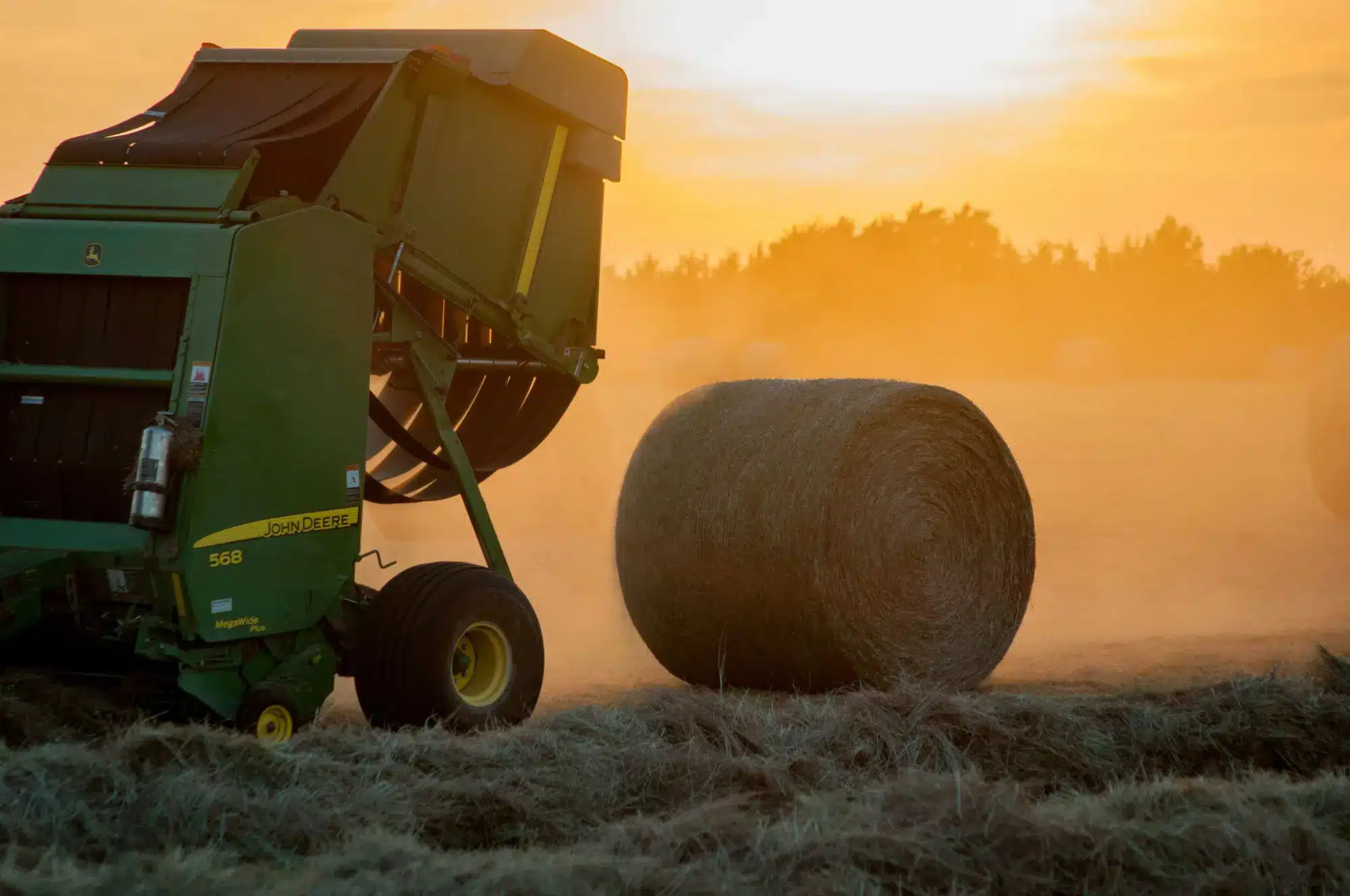 Round hay bale in a field, next to a bailer, with a sunrise behind.